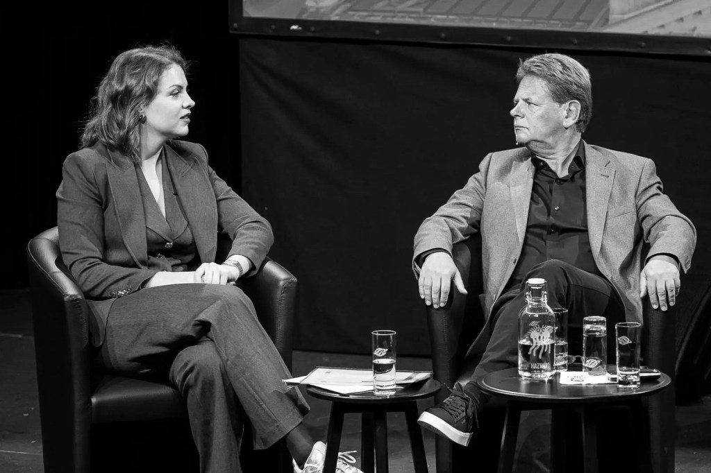 A black and white image showing two individuals seated in armchairs on stage. One person, wearing a suit, is engaged in conversation while the other, in a lighter-colored suit, listens attentively.