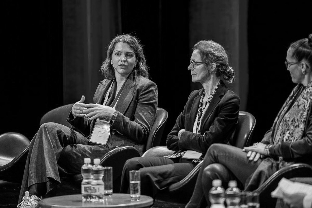 A panel discussion featuring three women, with one woman speaking passionately while the others listen attentively. The setting includes chairs and tables with water bottles, and the image is in black and white.