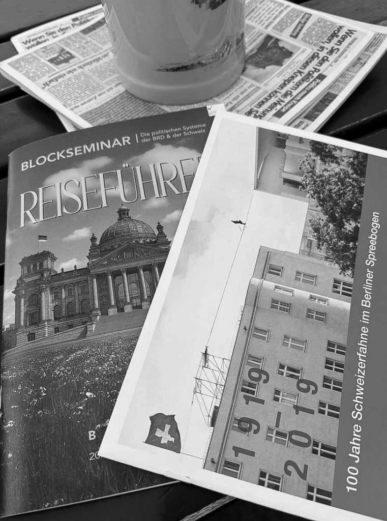A stack of printed materials on a table, including a travel guide titled 'BLOCKSEMINAR REISEFÜHRER' featuring the Berlin Reichstag building and a brochure about the 100th anniversary of the Swiss flag in Berlin, accompanied by a glass of beverage and newspapers.